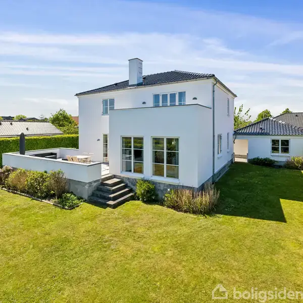 Hvidt hus med sort tag i moderne stil står på en velplejet græsplæne; terrassen findes på siden af huset, omgivet af hække og blomsterbede under en klar himmel.