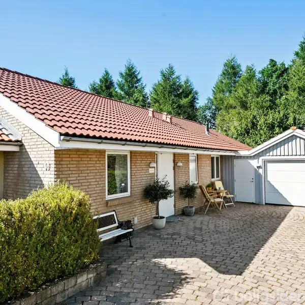 Et murstenshus med rødt tegltag har en lille gårdhave med flisebelægning. Ved siden af er en garage. Omkring huset ses grønne buske og træer under en klar blå himmel.