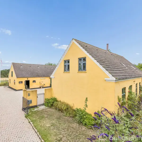 Gulmalet hus med stejlt tag og vinduer i tagetagen, står stille i en have med fliser foran; omgivet af buske og en blå himmel i baggrunden.