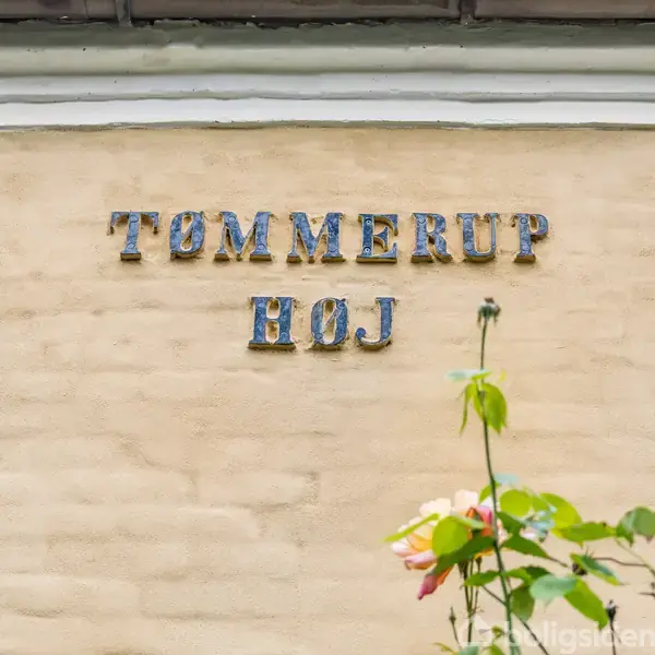 Tommerrup Høj-skilt er fastgjort til en lys murstensvæg. To roser blomstrer i forgrunden og tilføjer farve. Detaljerede murværk stiger til tagets kant.