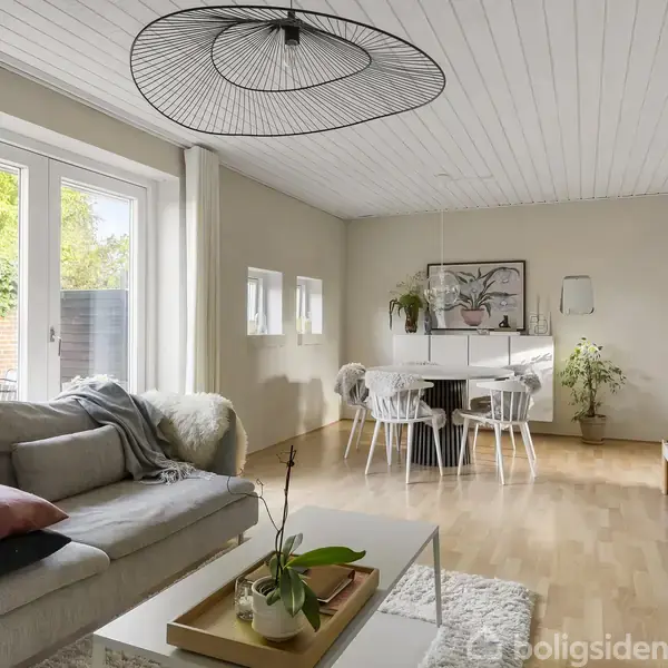En stue med en lys sofa og puder, står ved siden af et sofabord med planter. Rummet har store vinduer, trægulve og et spisebord med stole.