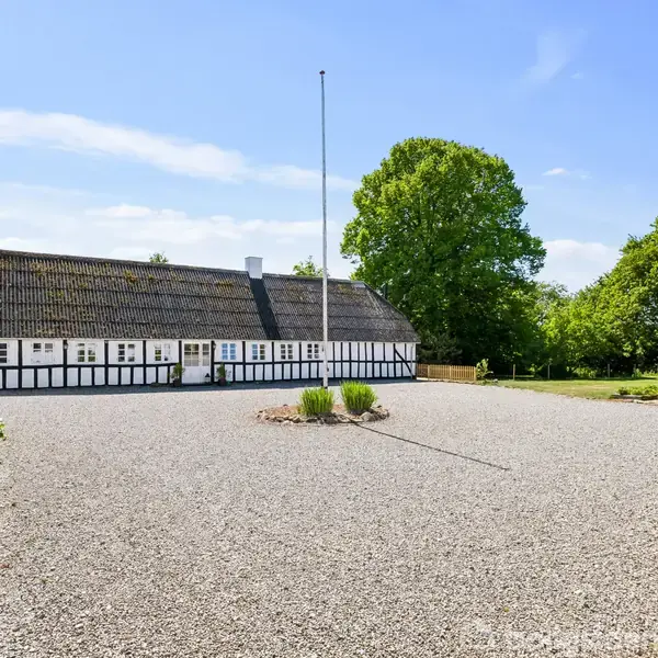 Bindingsværkshus står på en grusbelagt gårdsplads med et centralt placeret flagstang; køkkenhaverammer og drivhus findes i omgivelserne af grønne træer.