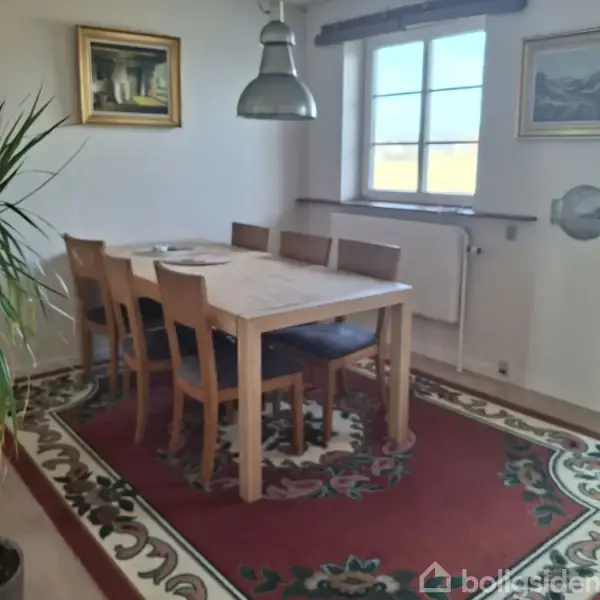 Et træbord med seks stole står på et mønstret tæppe i et rum med et vindue og radiator. Der hænger en lampe over bordet.