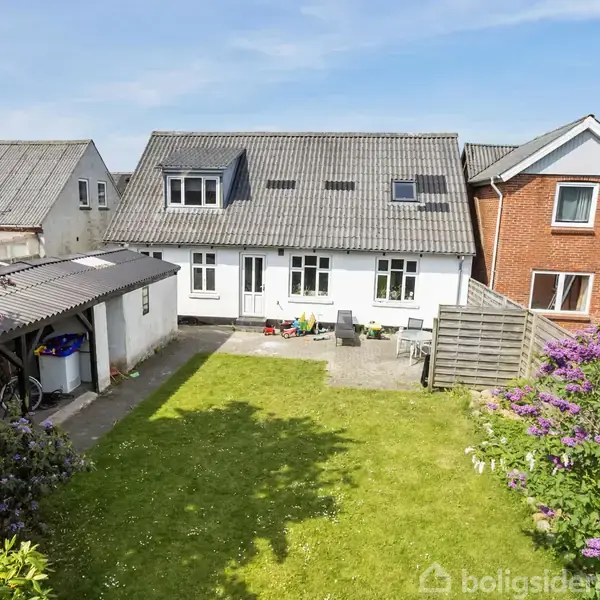 Huset med gråt tag står i midten, omkranset af en velplejet græsplæne og farverige blomster. Til venstre er der en carport fyldt med cykler. Blå himmel i baggrunden.
