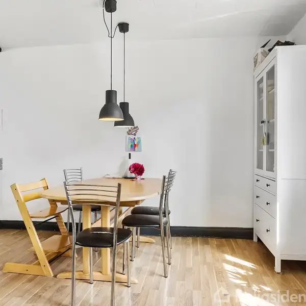 Et rundt spisebord står i en spisestue omgivet af fire stole. Over bordet hænger to sorte lamper. Rummet har hvide vægge, trægulv og et skab ved siden af vinduet.