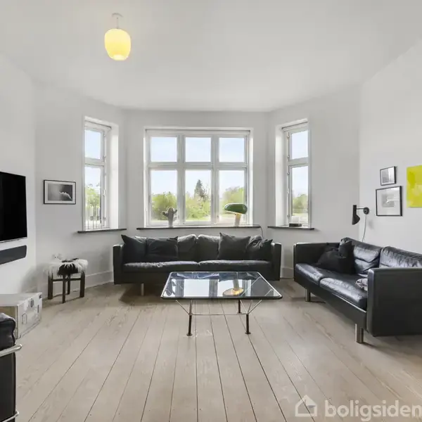 Sofaer arrangeret omkring et glasbord i en lys stue med trægulv, store vinduer og moderne kunst på væggene. En fladskærms-TV hænger ved siden af kunstværker.