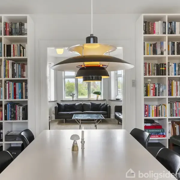 Et spisebord omgivet af sorte stole står i et rum med bogreoler fyldt med bøger. En moderne lampe hænger over bordet. I baggrunden ses en stue med en sort sofa.