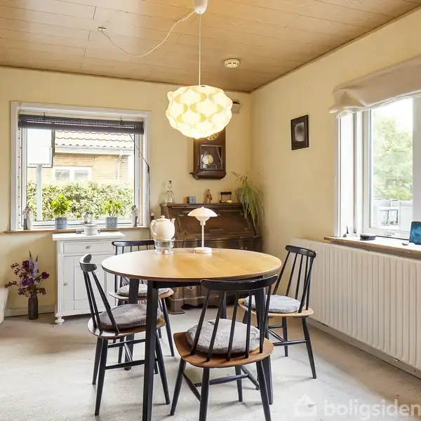 Et rundt spisebord med fire stole står centralt i et lyst køkken. Bordet har en lampe hængende over sig. Rummet indeholder vinduer, en radiator og pyntegenstande på bord og vindueskarm.