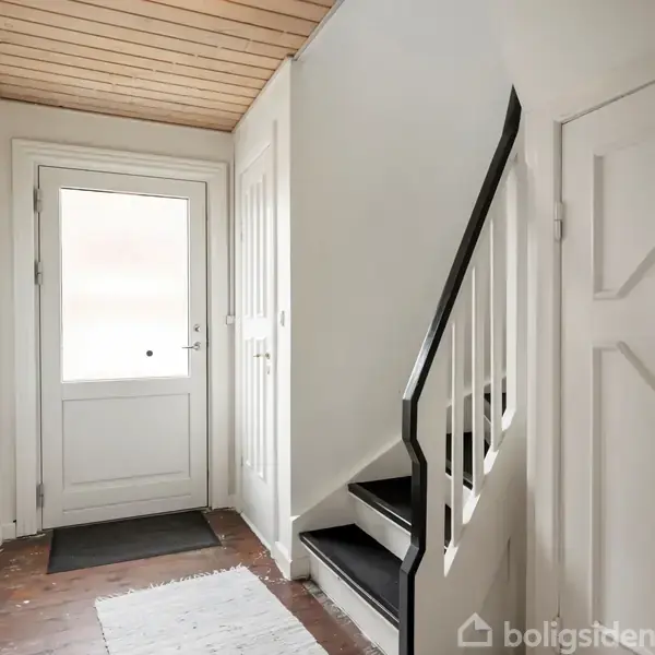 Dør fører ind til entré med trappe. Trappen går op til næste etage. Rummet har hvidt loft og vægge med brun trægulv og en dørmatte foran indgangsdøren.