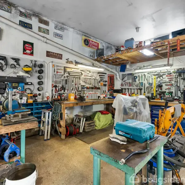 Et værksted fyldt med værktøj hænger på væggene. Arbejdsborde er dækket med forskellige genstande, som boremaskiner og værktøjskasser. Flere hylder opbevarer ekstra udstyr og materialer.