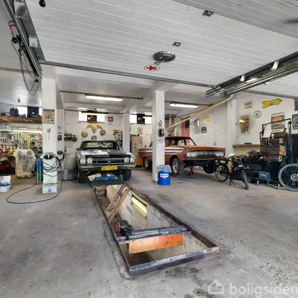 En autoværksted indeholder to biler, reparationsudstyr og en gulvgrav. Bilerne står parkeret, mens værktøj og cykler er opstillet rundt om. Lokalet er rummeligt og velorganiseret.