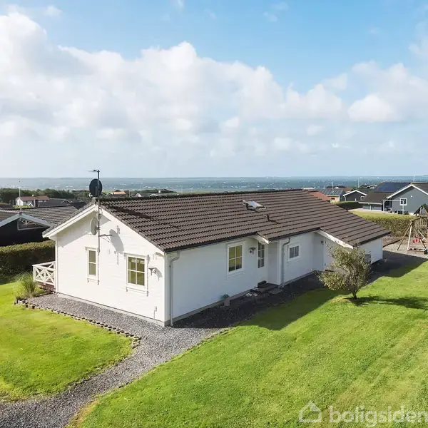 Hvidt hus med brun tag, står på en velplejet græsplæne omgivet af hække. Baggrund viser flere huse og en himmel med skyer.