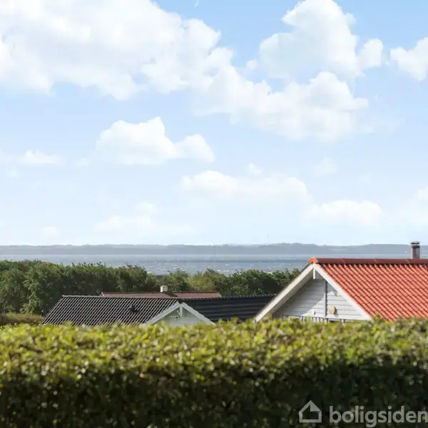 Huse med røde og sorte tage står stille bag en grøn hæk, med udsigt til blå himmel og hav samt skovklædte områder i baggrunden.
