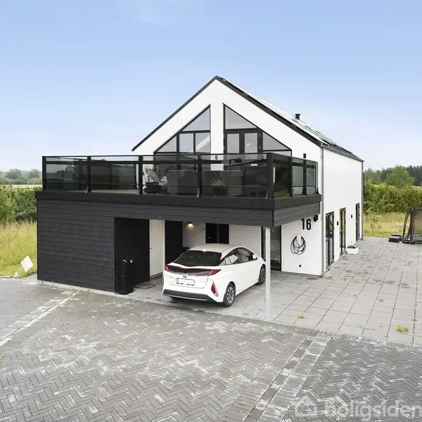 Et moderne hus i to etager med sort-hvide detaljer har en balkon på første sal. En bil er parkeret under balkonen. Husnummeret "16" er synligt på facaden. Græsmarker omgiver huset.