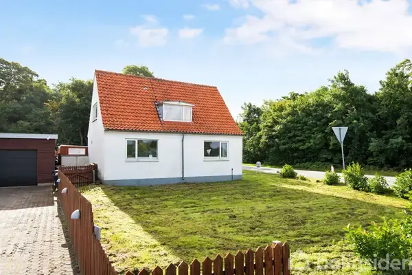 Huset står i en grøn have med et rødt tag og hvid facade. En lille sti omkranset af et lavt hegn fører til hoveddøren. Skovklædt område i baggrunden.