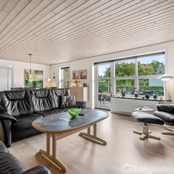 Sort lædermøblement står stillet i en stue. Sofasæt og lænestol omkranser et ovalt sofabord med en grøn skål. Store vinduer tilbyder udsigt til en grøn have.