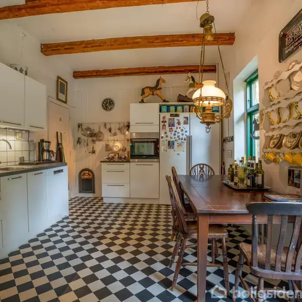 Køkken med skakternet gulv og rustikke træbjælker. Hvide skabe og fliser, ovn og køleskab foran spisebordet med fire stole. Dekorationer på væggen og hængende lampe.
