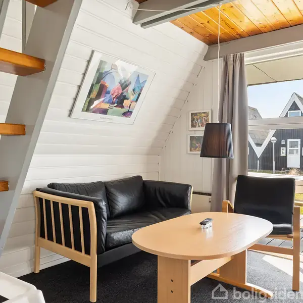 Stue med sort lædersofa, rundt træbord og stigelignende trappe. Store vinduer viser udsigt til terrasse med moderne huse i baggrunden. En lampe hænger over bordet. Maleri på væggen.