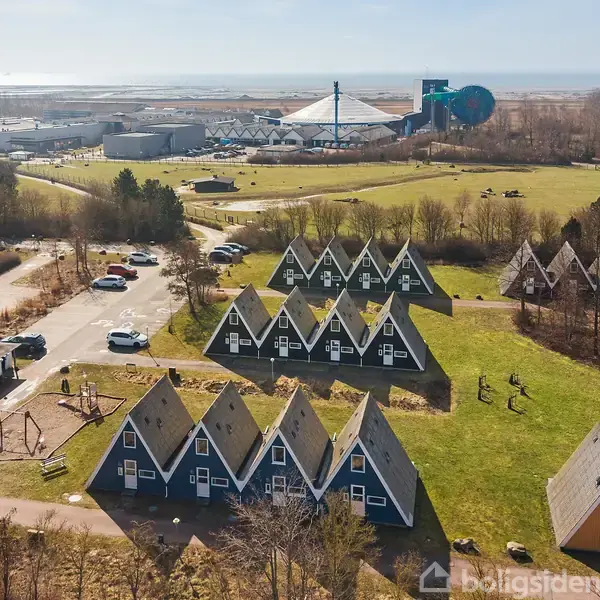 A-formede huse står stille i en række på en grøn mark. I baggrunden ses et større bygningskompleks, og biler holder parkeret langs en vej mellem husene.