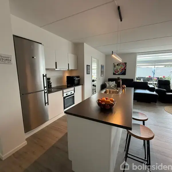 Moderne køkken med køleskab, ovn og frugtskål på køkkenø. Stole står ved øen, og en lys stue med sofa og vindue ses i baggrunden. Skilt: "Mommy’s KITCHEN".