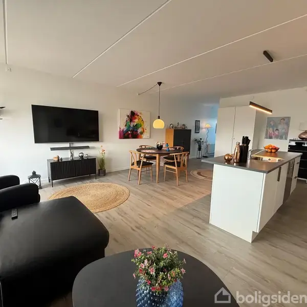 En moderne stue med sort lædersofa, fladskærms-TV på væggen, og en åben køkkenø. Et spisebord med stole står ved siden af. Lyse trælaminatgulve og minimalistisk indretning omkranser rummet.