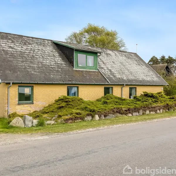 Et gulmurstenshus med skråtag ligger ved en vej. Huset er omgivet af buske og træer i baggrunden, og har grønne vinduesrammer. Der er en asfalteret vej foran.