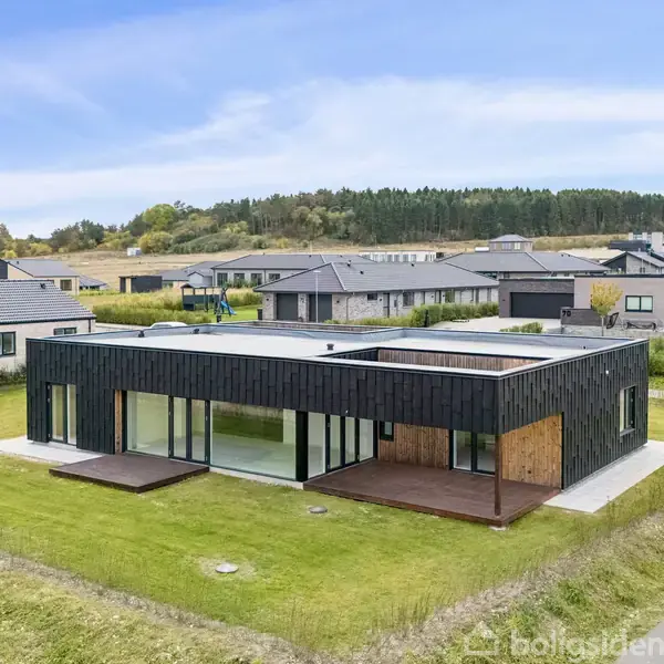 En moderne, kubisk bygning med sorte paneler står på en velplejet græsplæne. Omkringliggende område består af andre moderne huse og en skov i baggrunden under en blå, let skyet himmel.