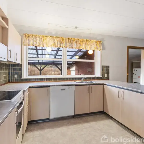 Køkken med lyse træskabe og grå bordplader, i brug med opvaskemaskine og ovn. Store vinduer med blomstrede gardiner giver udsigt til overdækket terrasse.