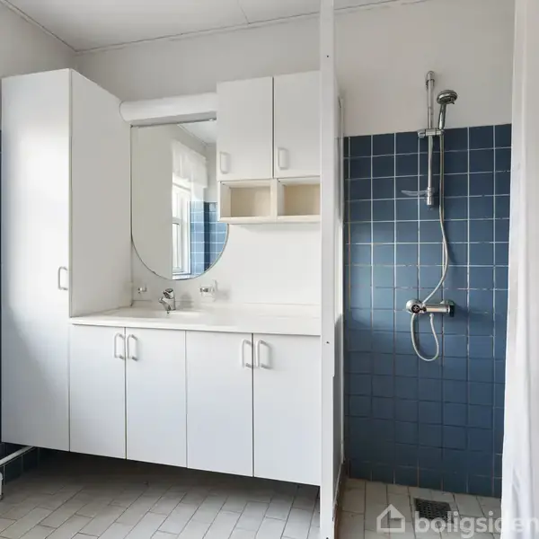 Hvidt badeværelseskab står mod en væg, med et rundt spejl over vasken. Bruseren er på en væg af blå fliser. Vindue med gardin giver naturligt lys.