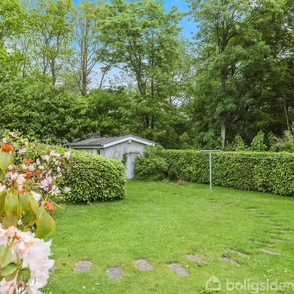 Haven med græsplæne er omgivet af frodige buske og træer. En lille grå skur står i baggrunden. Foran er blomstrende planter med lyserøde og orange blomster.