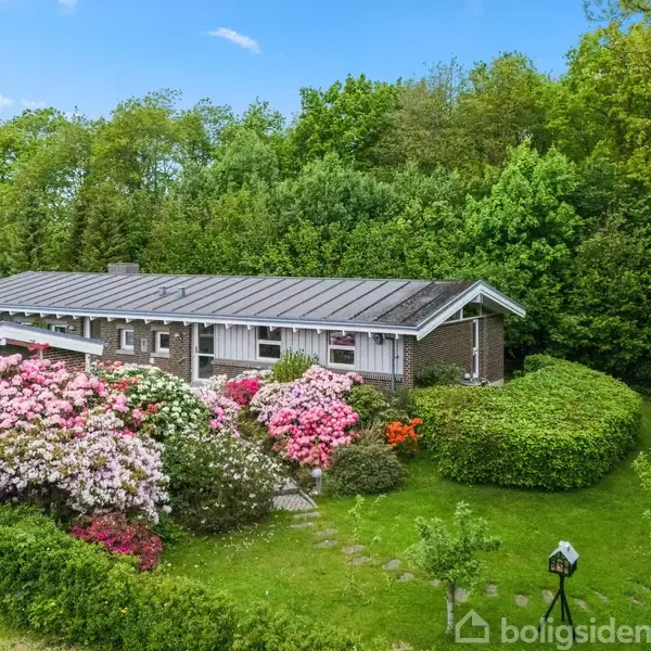 Huset er omgivet af farverige blomsterbede og tæt bevoksning. Det har et fladt tag, brune murstensvægge og er placeret i en grøn, frodig have med buske og træer.