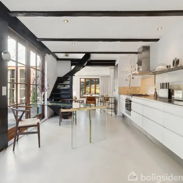 Et moderne køkken med hvide skabe og køkkenudstyr arrangeret langs væggen. En glasbord står centralt, omgivet af stole. Store vinduer og en dør fører ud til en terrasse.