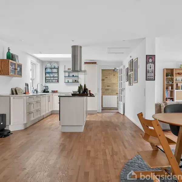 Et moderne køkken med træbordplade på en central ø. Køkkenet er en del af et åbent rum med spisebord og stole på trægulv. Reoler i baggrunden.