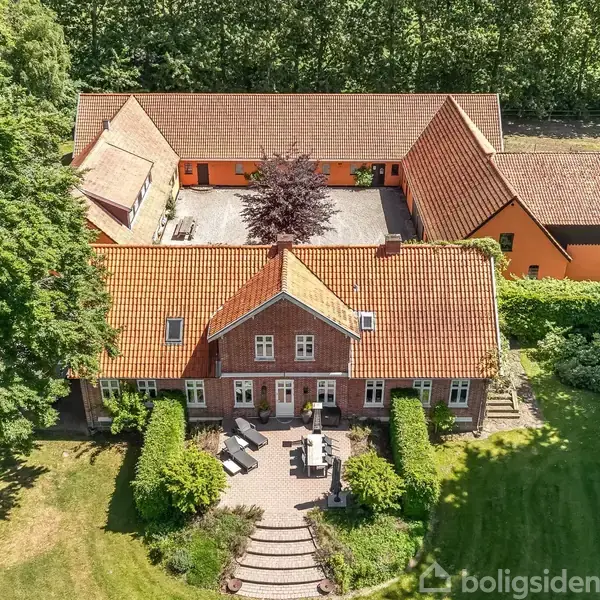 Hus med rødt tegltag omgivet af grønne træer og have. Indre gårdsplads med flere bygninger rundt om. Terrassemøbler er placeret foran huset. En symmetrisk sti fører op til hoveddøren.