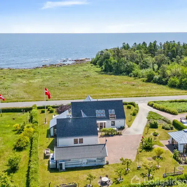 Et hus med en mørkeblå tag, flankeret af en grøn have, står stille. I baggrunden er der en vej, græsarealer og en kystlinje med havudsigt. To danske flag er hejst.