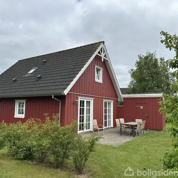 Et rødt træhus står stille i en grøn have med græs og buske. Ved siden af huset ses en terrasse med havemøbler. Himlen er overskyet.