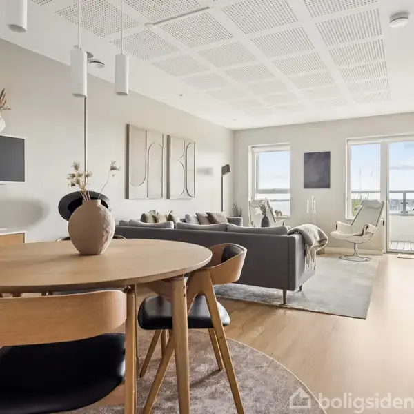 Et rundt træbord med stole står i en moderne stue, hvor en sofa og lænestole vender mod store vinduer, der giver udsigt til en balkon og lysindfald.
