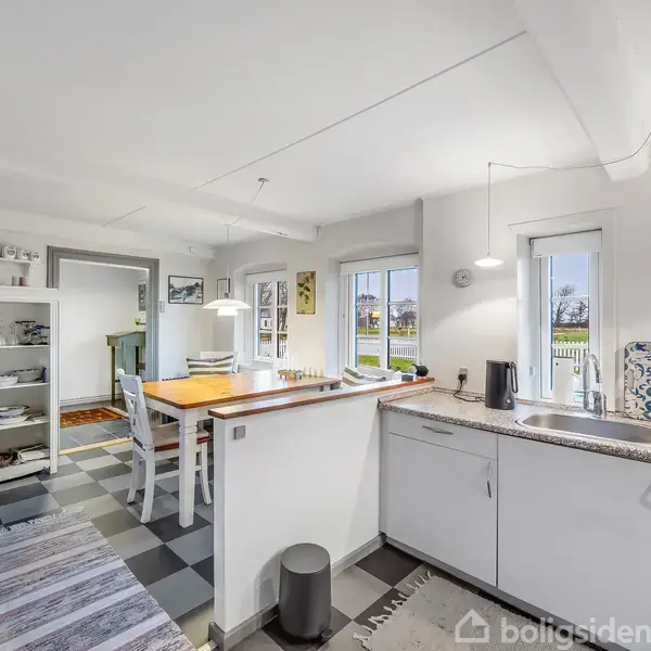 Køkken med hvidlakerede skabe står stille og skinnende i et lyst rum med store vinduer. Et spisebord med stole står centralt. Kurver og tæpper dekorerer gulvet.