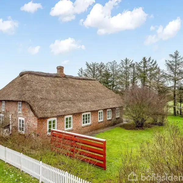 Et bindingsværkshus med stråtag står stille omgivet af en grøn have med buske og træer. Hvide og røde hegn omkranser ejendommen. Himlen er blå med skyer.