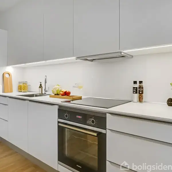 Et moderne køkken med hvide skabe og en indbygningsovn. Spisebordet er beliggende uden for rummet. På bordpladen står krydderier, en plante og tilberedningsvarer.