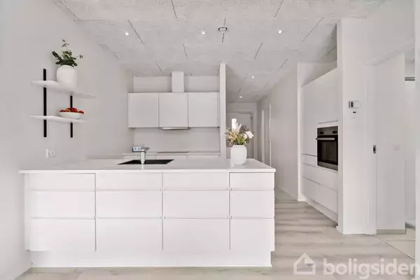 Et moderne køkken med hvidt interiør; skabsø i forgrunden, komfur og vask på toppen. To hylder med frugter og planter til venstre. Indbygningsovn til højre.