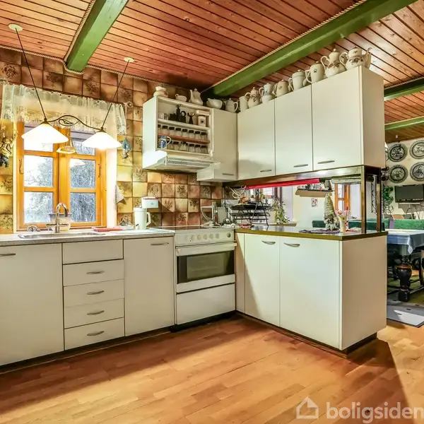 Et køkken har hvide skabe og skuffer, en komfur og en emhætte. Det er lyst op af vinduer med gardiner. Tredimensionale fliser på væggene. Rummet har træloft og gulv.