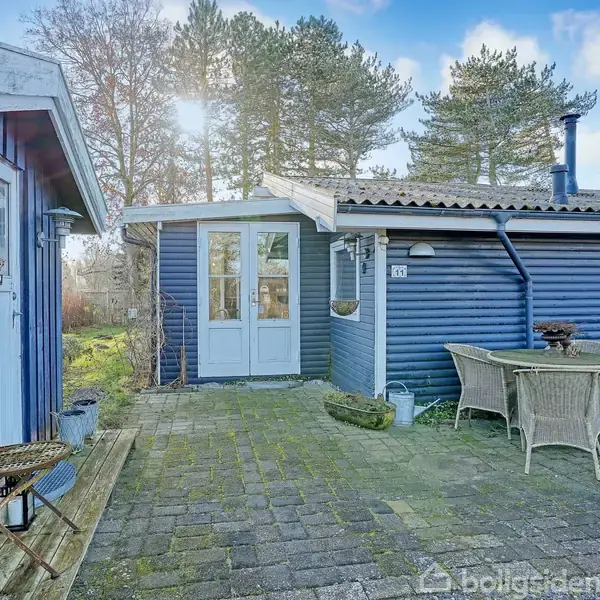 Blåt træhus med fladt tag og dobbeltdør står i en brolagt gårdhave. Ved siden af står et rundt bord med fire kurvestole. Træer og himmel i baggrunden.