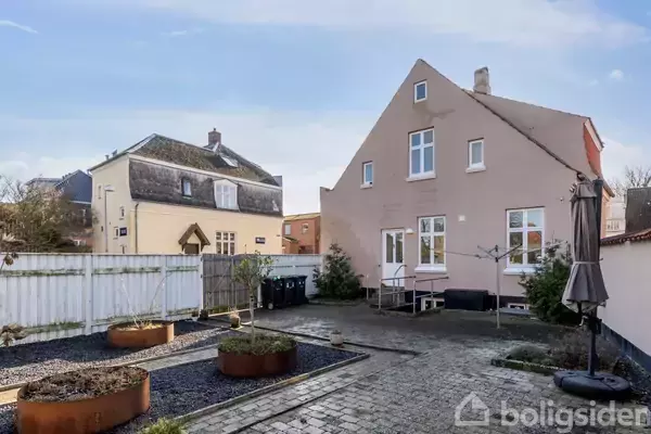 Hus med skråt tag står i en brolagt gårdhave, omgivet af højbede og havekrukke i metal. Nabohus ses i baggrunden under en blå himmel. Ingen tekst til stede.