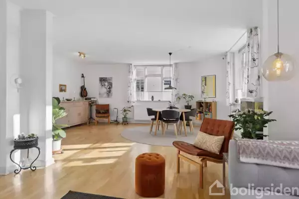 En moderne stue med trægulv indeholder et spisebord med stole. En lampe hænger over bordet. Rummet er lyst med vinduer, planter og en guitar på væggen.