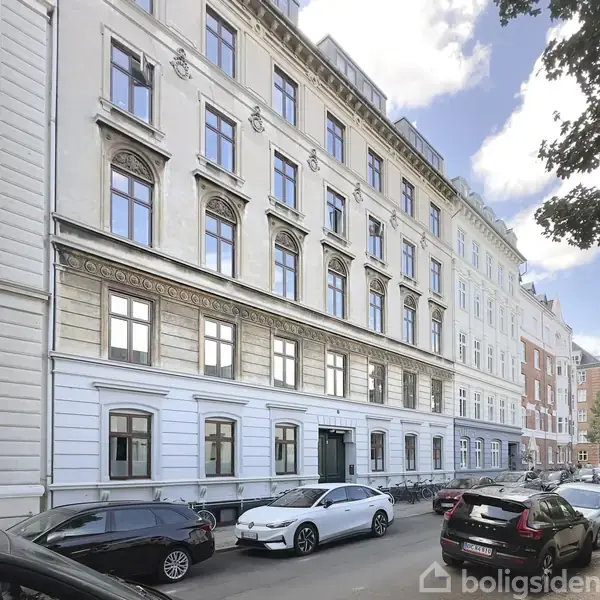 Bygning med flere etager står på en byvej. Biler er parkeret langs gaden foran bygningen, som har store vinduer og en klassisk facade. Træer ses til højre.