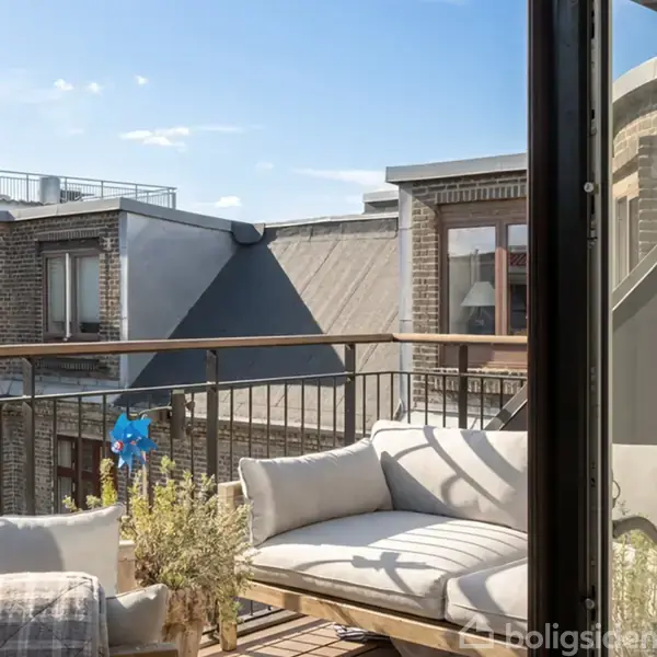 Blød sofa står på en balkon, med udsigt til bygningens murstensfacade og skråtage under en klar blå himmel.