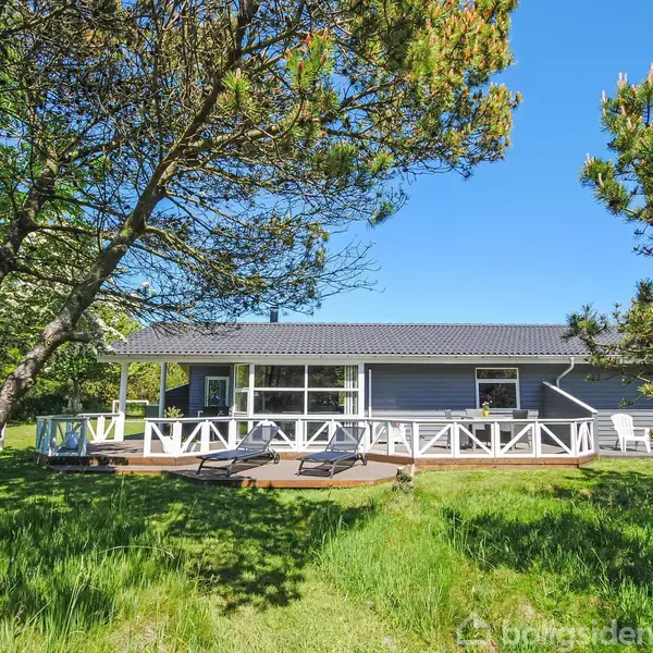 Et sommerhus med træterrasse ligger omkranset af grønne træer og en græsplæne under en klar, blå himmel. Der er liggestole og et udendørs bord og stole på terrassen.