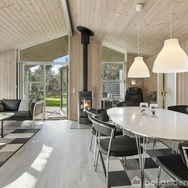 Stuen indeholder en sort brændeovn ved væggen, omgivet af en spisestue med hvidt bord og sorte stole. Store vinduer og døre åbner ud til en grøn have.