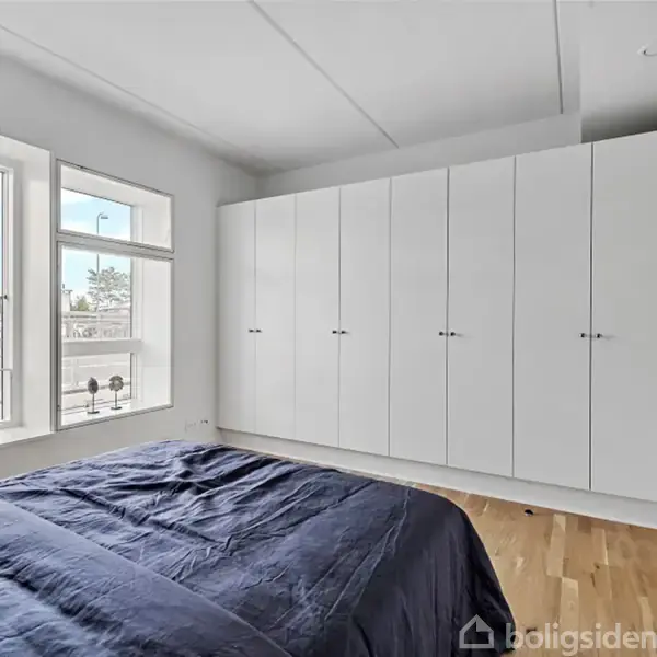 Soveværelse med en stor indbygget hvid garderobe, placeret langs væggen. En seng med mørkeblå sengetøj står foran store vinduer, der giver naturligt lys med udsigt over gaden.